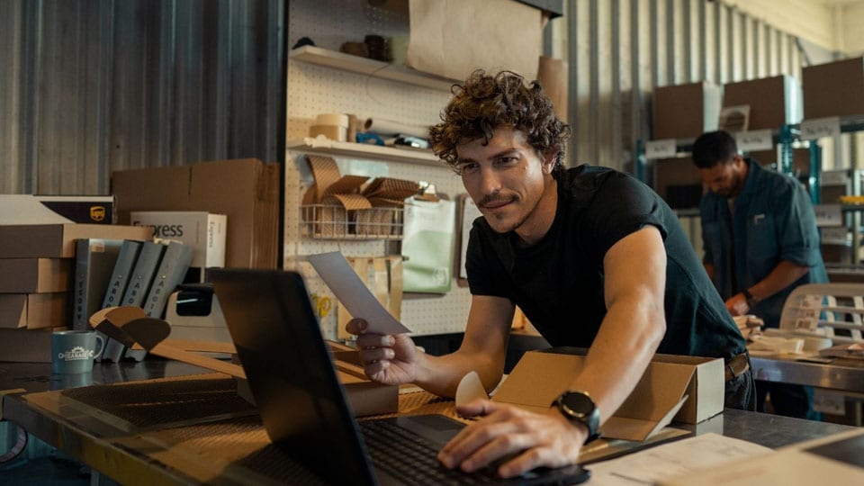 man in workshop leaning on desk looking at open laptop